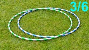 Two rings lying on the grass. The rings are the same shape and their surfaces are coated with a shiny, glittery material. One ring has alternating white and green stripes. The other has alternating white and blue stripes. 3/6 is written at the top right of the image.